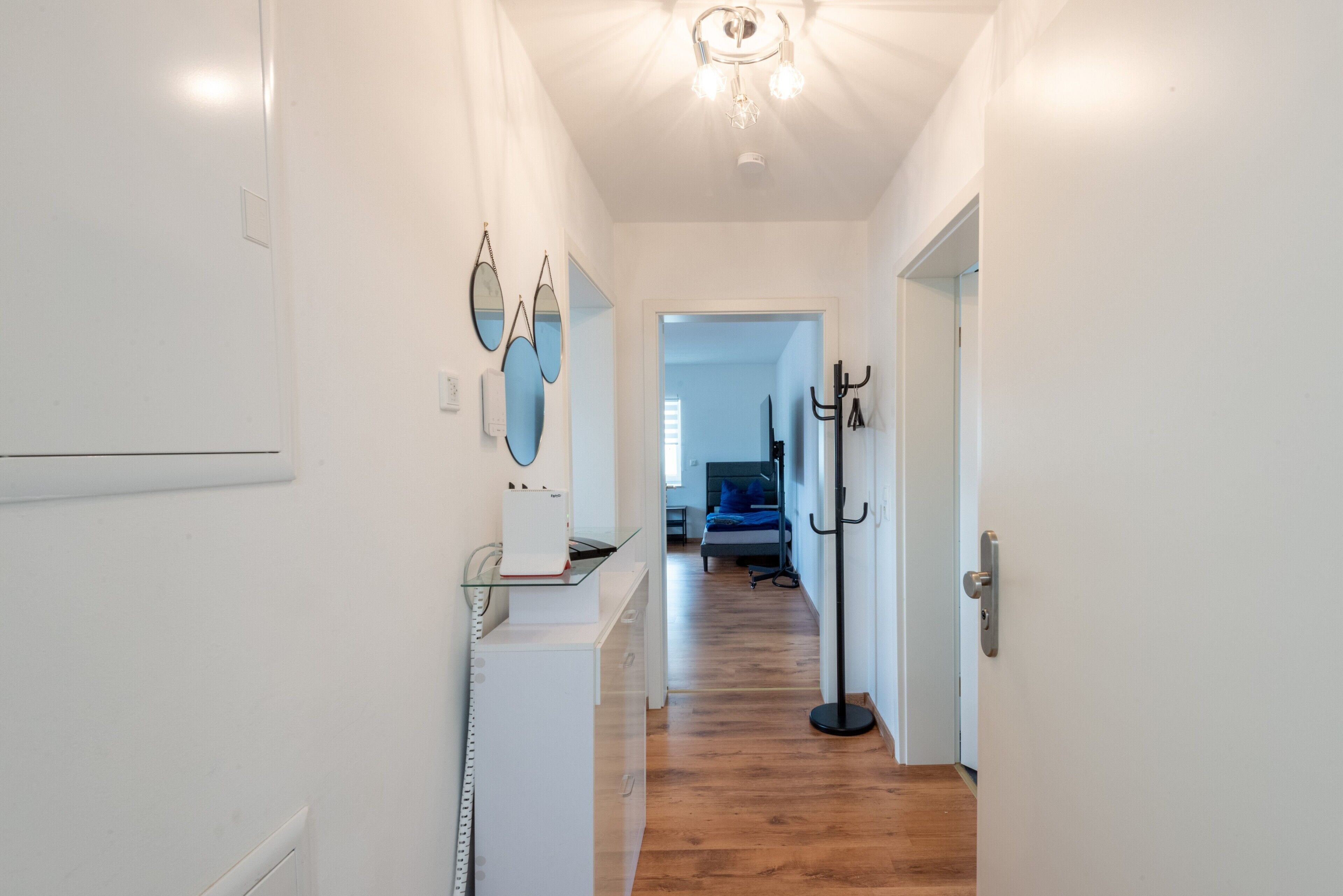A hallway with a wooden floor, white walls, a ceiling light fixture, and a bed with a blue headboard in the distance.