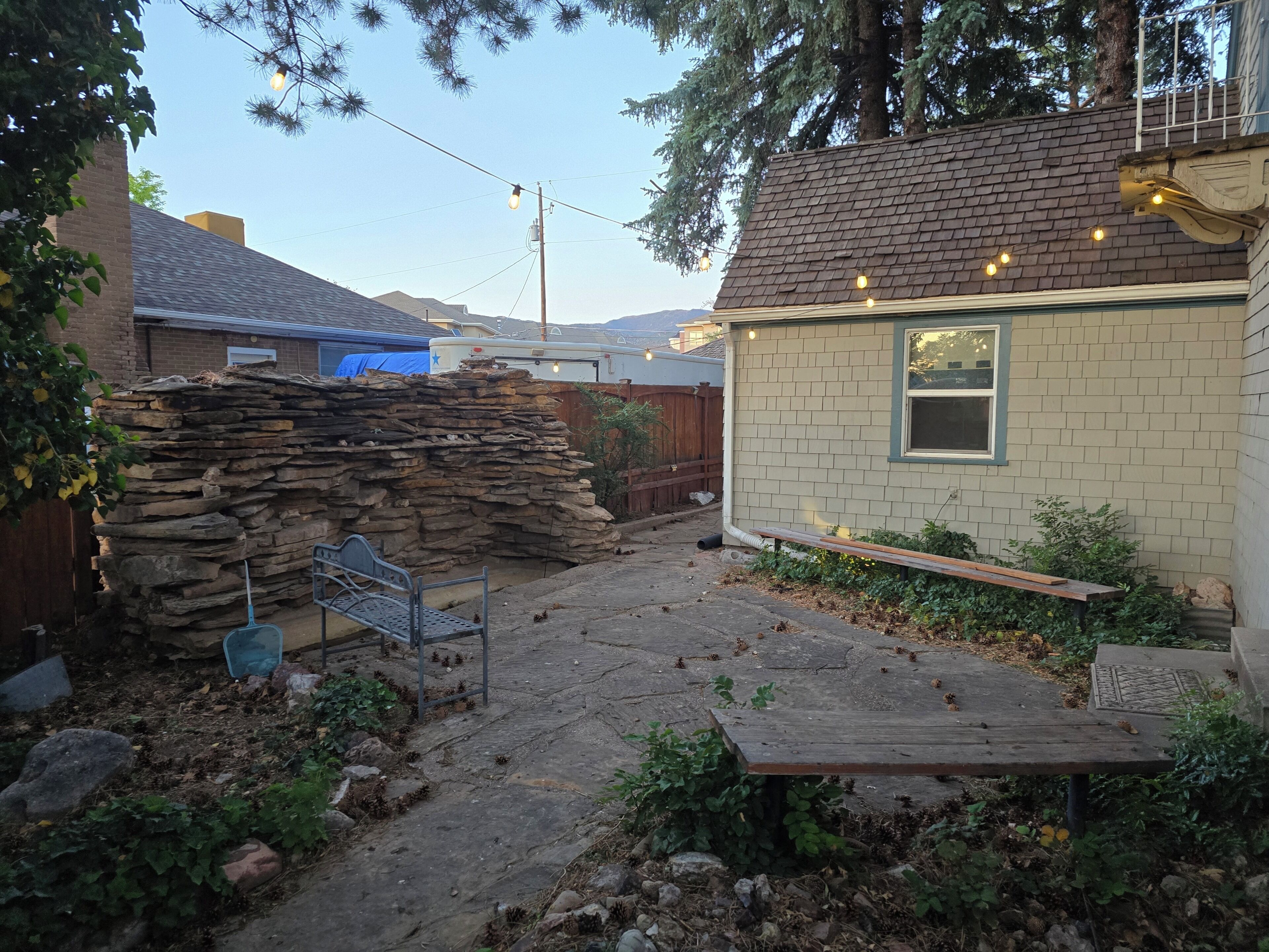 Un jardin avec un mur en pierre, un banc en métal, une table en bois et une maison avec une fenêtre.