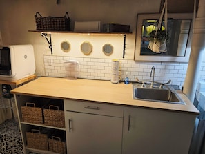 A kitchen with a white tiled backsplash, a stainless steel sink, a microwave, and a coffee maker. There are wicker baskets on the lower shelf and a hanging plant above the sink.