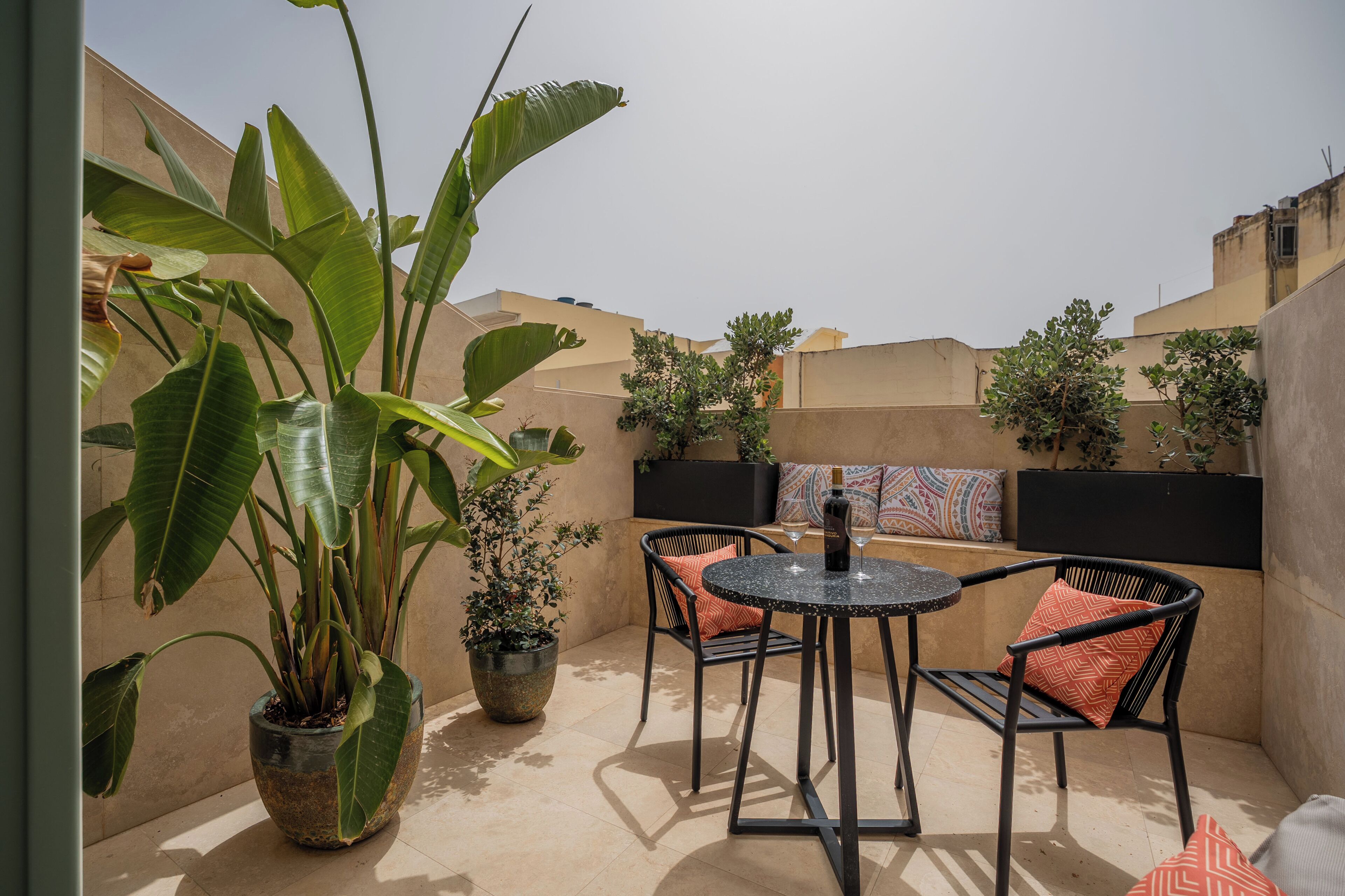 A patio with a round table and chairs, surrounded by potted plants and a view of buildings.