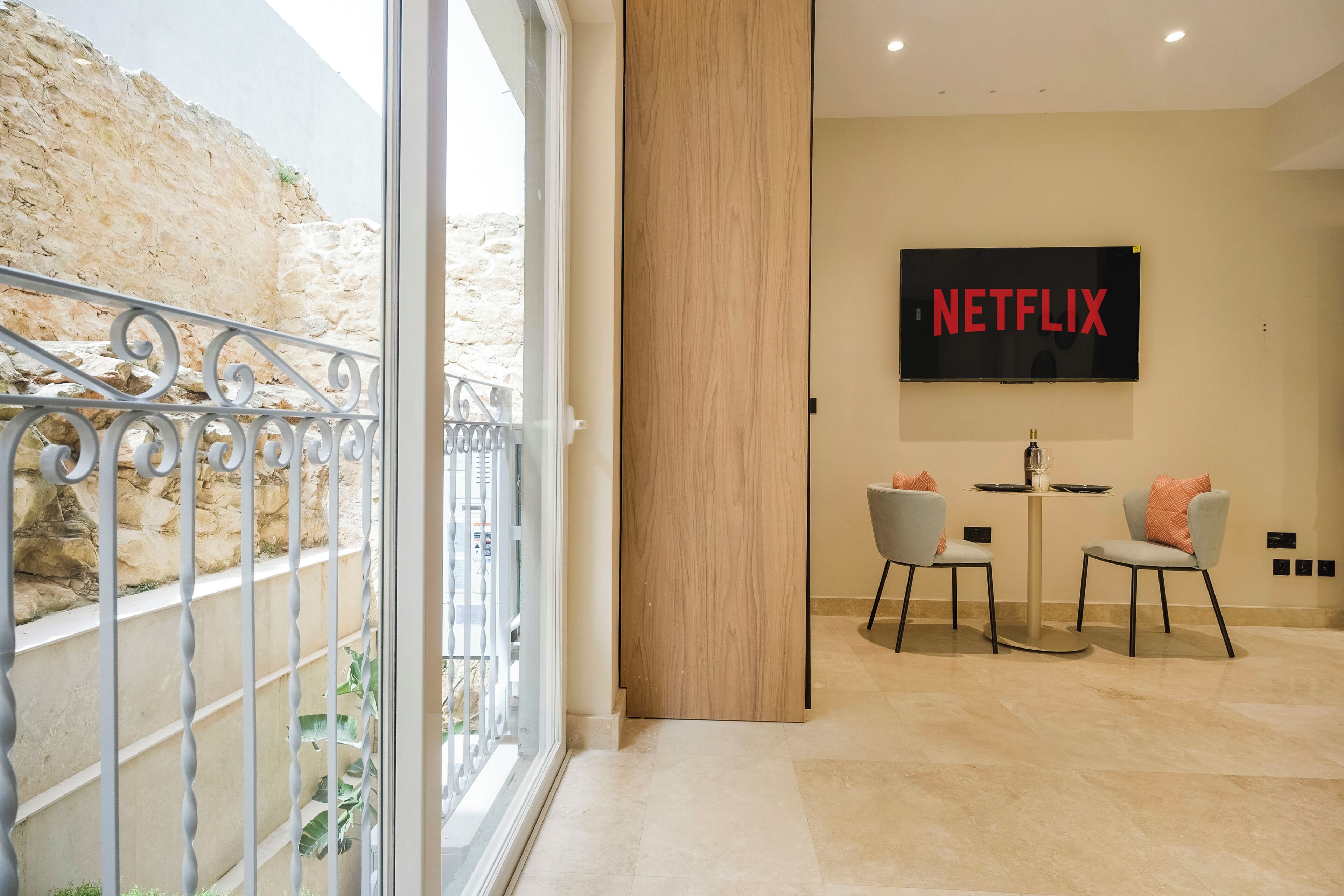 Un interior moderno con una gran pantalla de televisión mostrando 'NETFLIX', un comedor con dos sillas y un balcón con barandillas de metal.