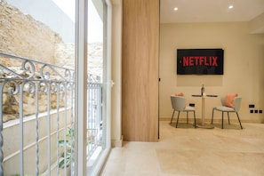Et moderne interiør med et stort fjernsyn, der viser 'NETFLIX', et spiseområde med to stole, og en balkon med gelændere i metal.