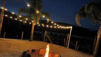 Escena nocturna al aire libre con un brasero, luces colgantes y palmeras.