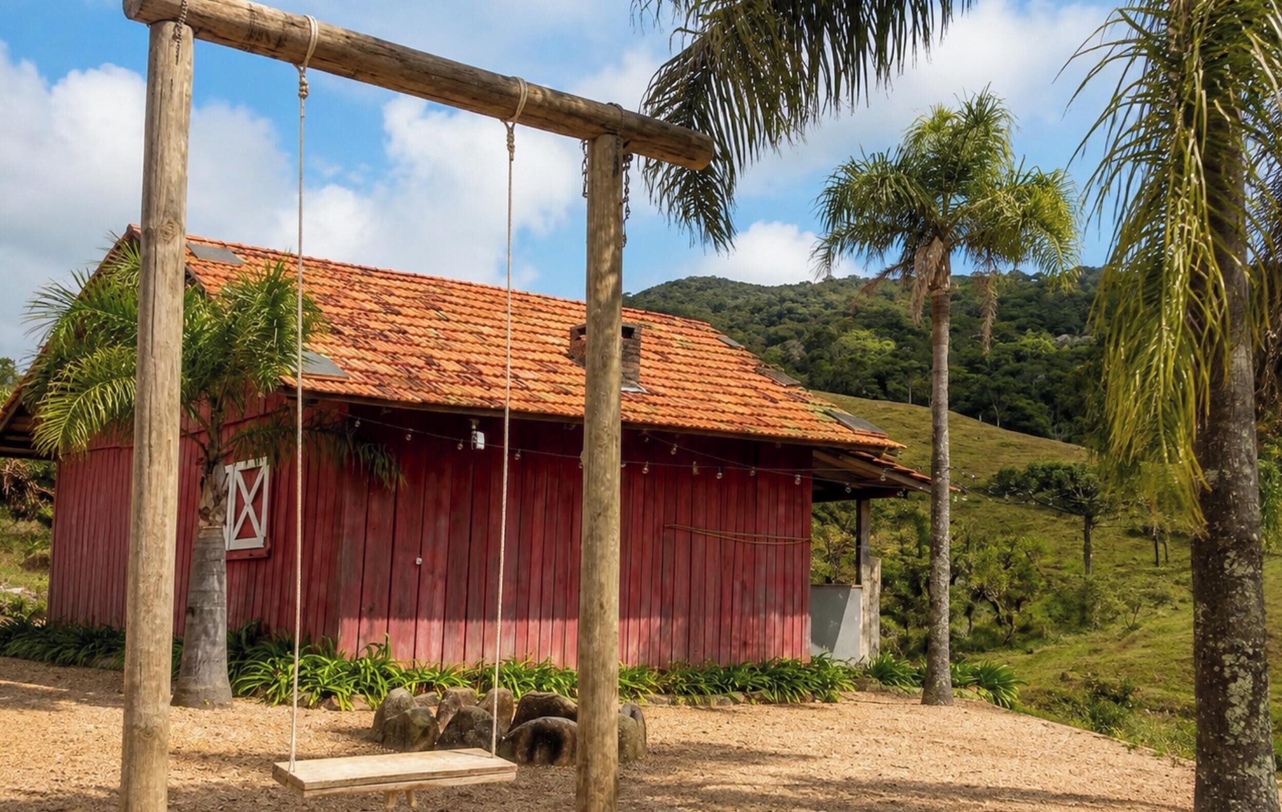 Un bâtiment en bois rouge, surmonté d’un toit en tuiles, entouré de palmiers et situé dans un paysage vallonné.