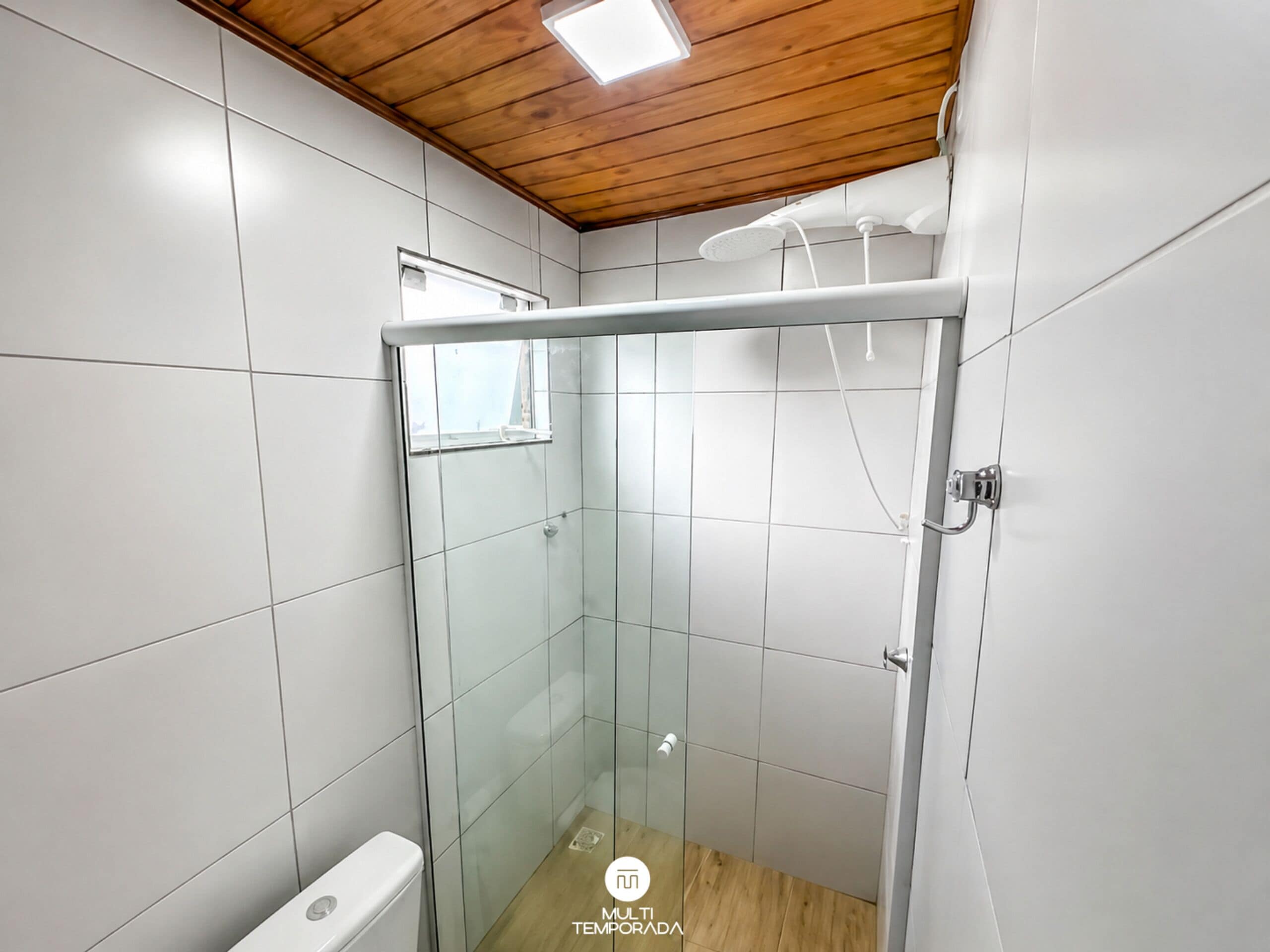 Pequeño espacio de ducha cerrado con puerta de cristal, paredes de azulejos blancos y techo de madera.