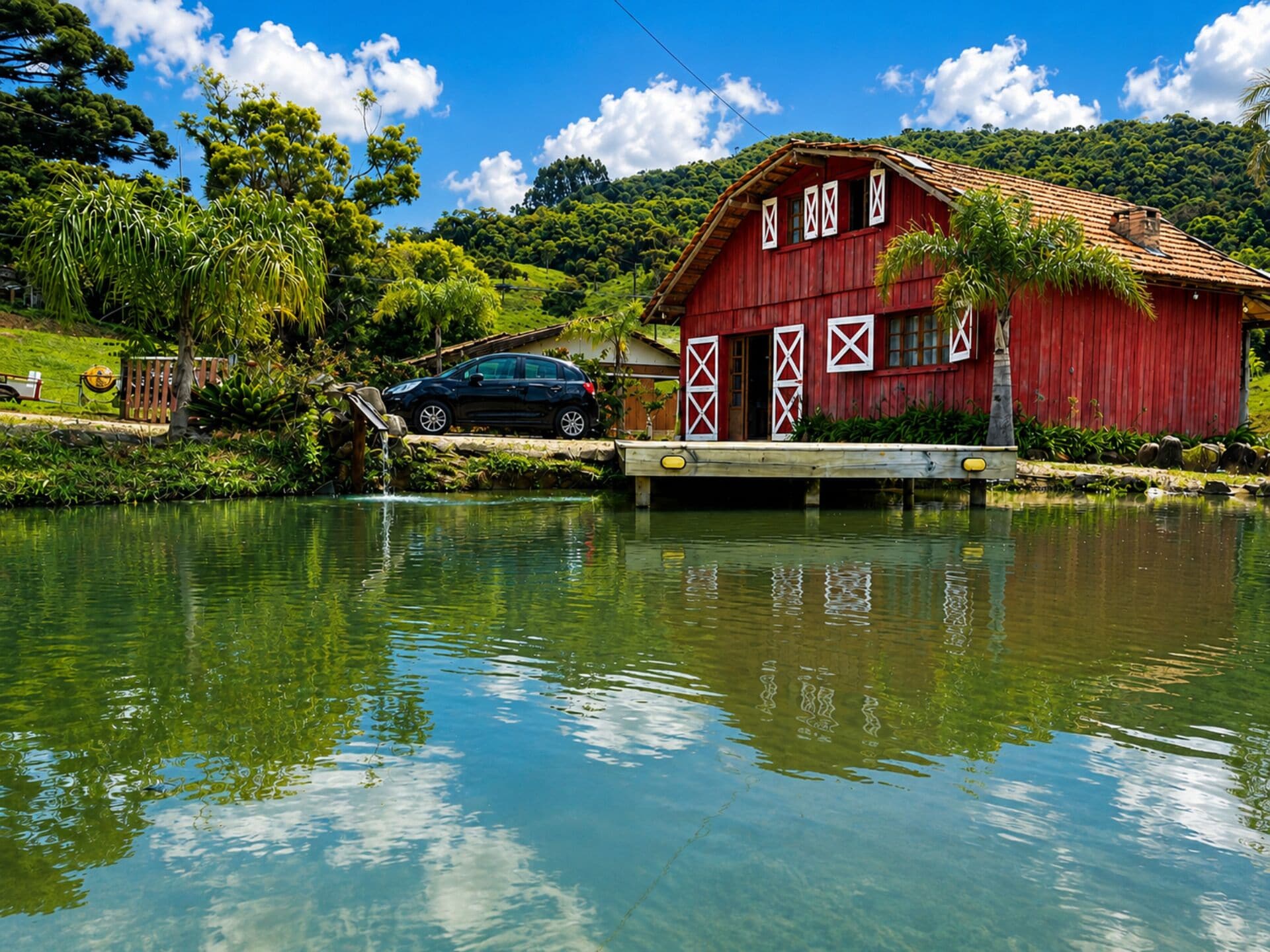 A red barn with white cross patterns, a car parked in front, and a calm lake reflecting the sky.