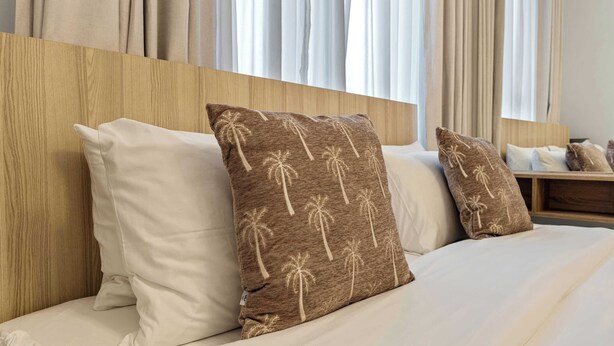 Hotellihuone, jossa on puinen pääty, valkoiset vuodevaatteet ja kaksi koristetyynyä, joissa on palmujen kuvioita.