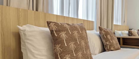 Habitación de hotel con cabecera de madera, ropa de cama blanca y dos almohadas decorativas con estampado de palmeras.