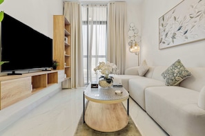 A modern living room with a flat-screen TV, wooden shelving, a round coffee table, a sofa, and a decorative wall piece. - 1BR Retreat near Expo City with Pool, Gym & Sports Courts (Jebel Ali)