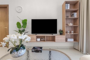 A modern living room with a flat-screen TV, a bookshelf, a clock, and a coffee table with decorative items. - 1BR Retreat near Expo City with Pool, Gym & Sports Courts (Jebel Ali)