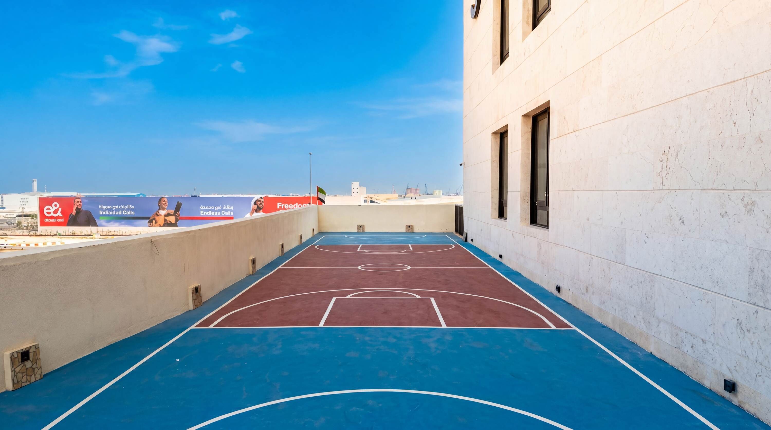 Een basketbalveld buiten met een blauw en rood oppervlak, naast een gebouw met witte muren en ramen.