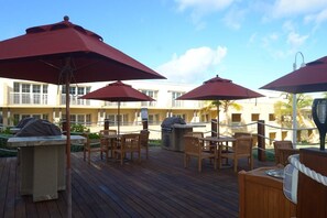 A wooden deck with outdoor seating and red umbrellas, a barbecue grill, and a poolside area with a building in the background. - 3 bedroom apartment at Dawn Beach Club, JW Marriott Resort, Beach and Spa (Oyster Pond)