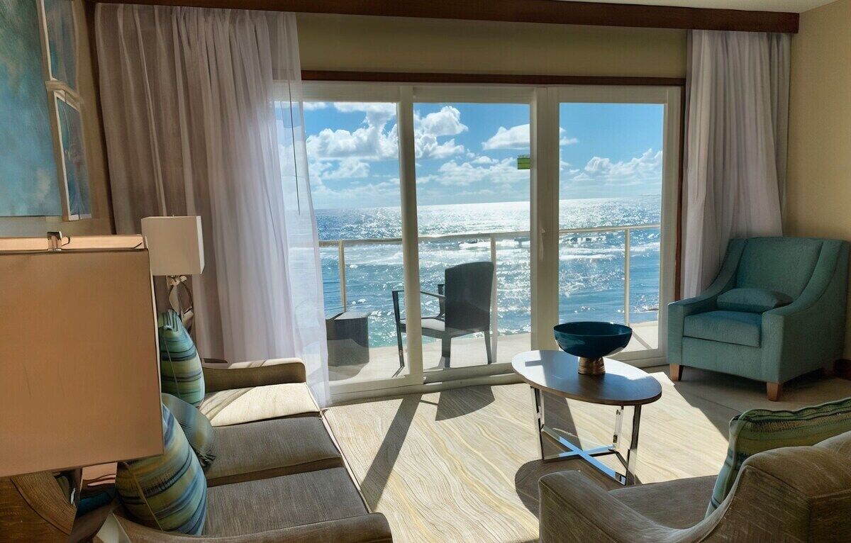 A room with a view of the ocean, a balcony with a table and chairs, and a seating area with a sofa and armchairs.