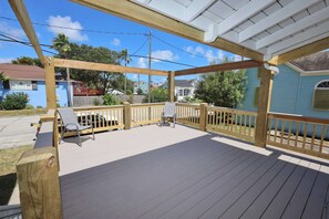 A covered outdoor deck with a wooden railing, a single chair, and a view of neighboring houses. - Cotton Cottage - Cozy Galveston Getaway (Galveston)