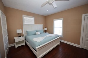 Une chambre à coucher comprenant un lit, deux tables de chevet, un ventilateur de plafond et deux fenêtres équipées de stores.