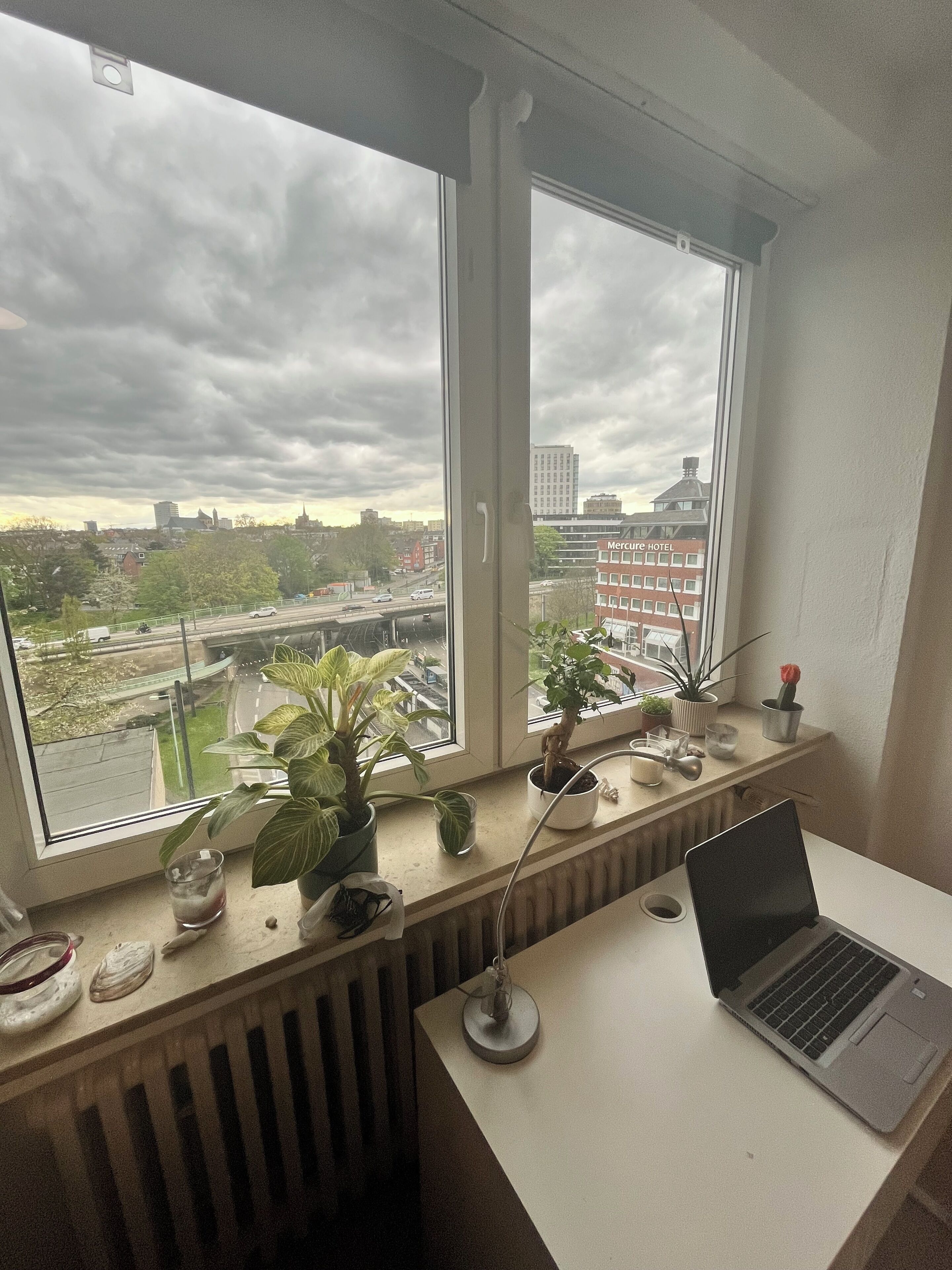 A room with a desk, a laptop, a radiator, and a window overlooking a cityscape.