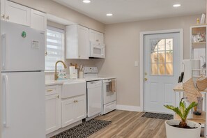Cocina con armarios blancos, microondas, lavavajillas y estufa. Hay una puerta con ventana, una planta y una encimera con utensilios de cocina.
