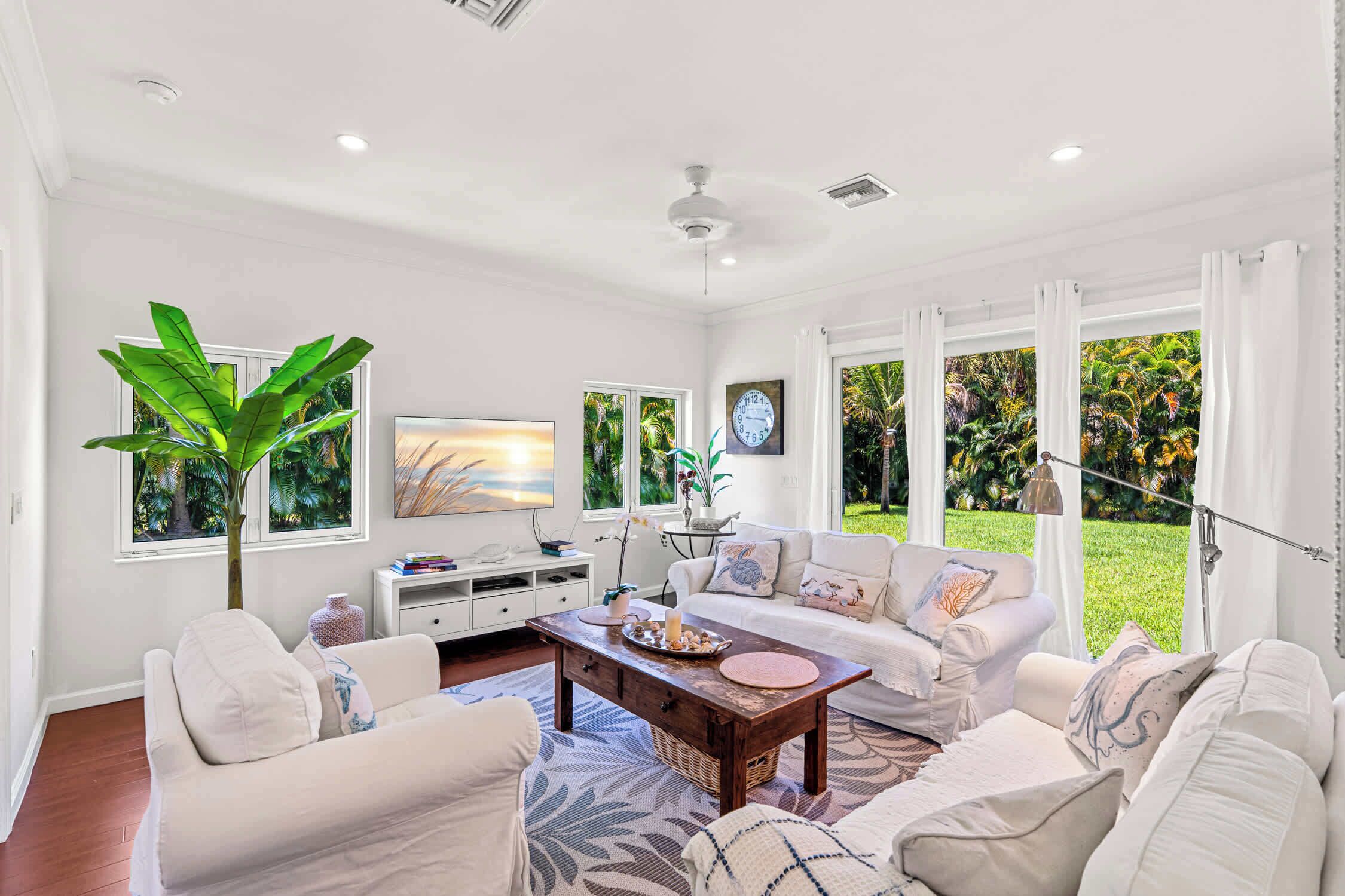A spacious living room with a large window, a ceiling fan, and a white sofa set.
