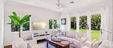 A spacious living room with a large window, a ceiling fan, and a white sofa set.