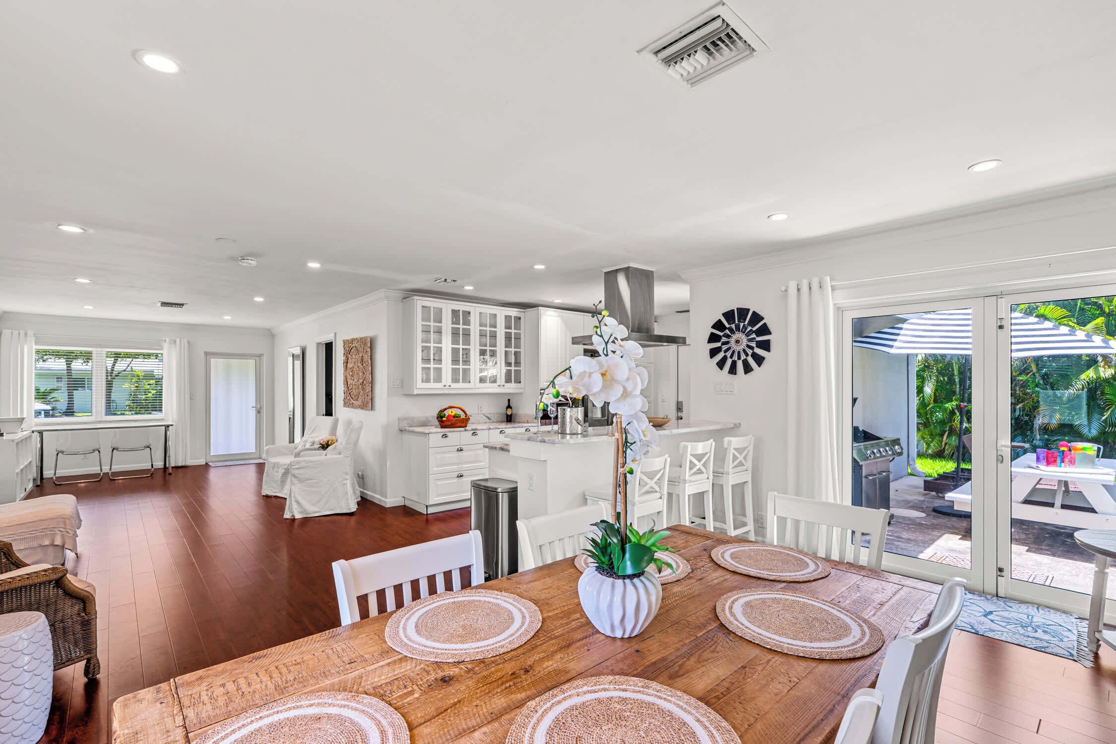 Una cocina amplia con armarios blancos y una isla central, un comedor con mesa de madera y individuales tejidos, y vista a un patio con mobiliario exterior.