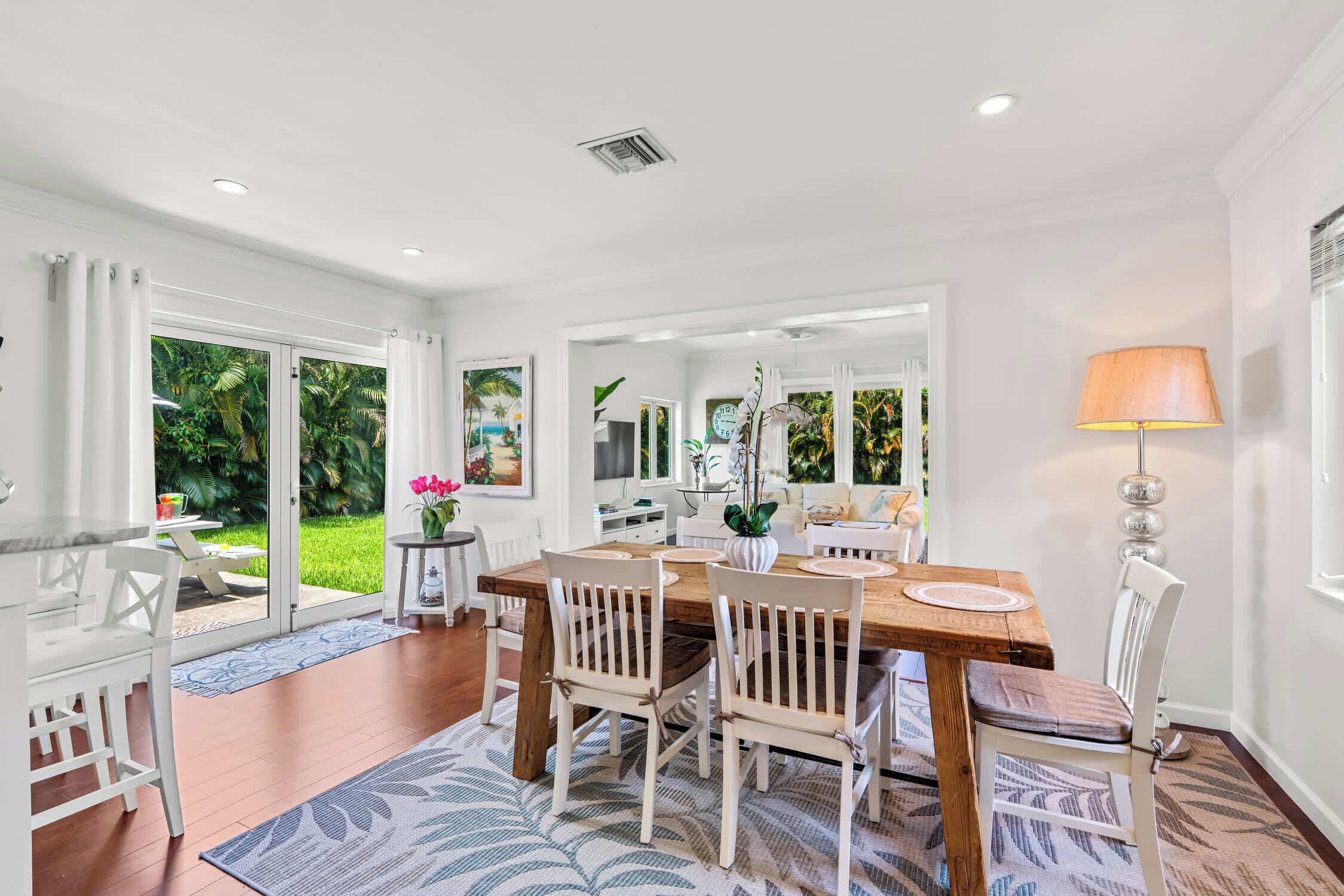 A spacious dining area with a wooden table and white chairs, a large window with a view of greenery, and a ceiling with recessed lighting.