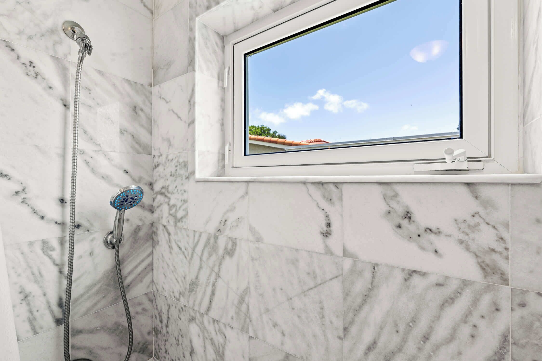Une salle de bain avec douche, une fenêtre donnant sur un bâtiment aux toits rouges et des murs en marbre.