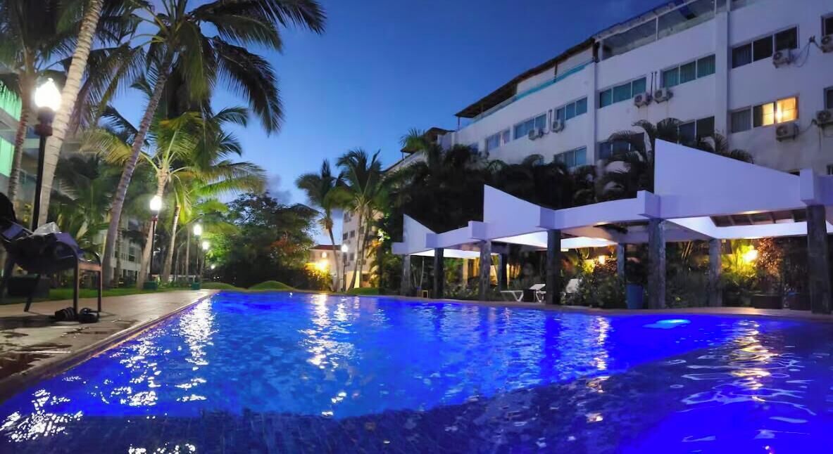 A swimming pool with a blue-lit edge, surrounded by palm trees and a building with multiple floors and balconies.