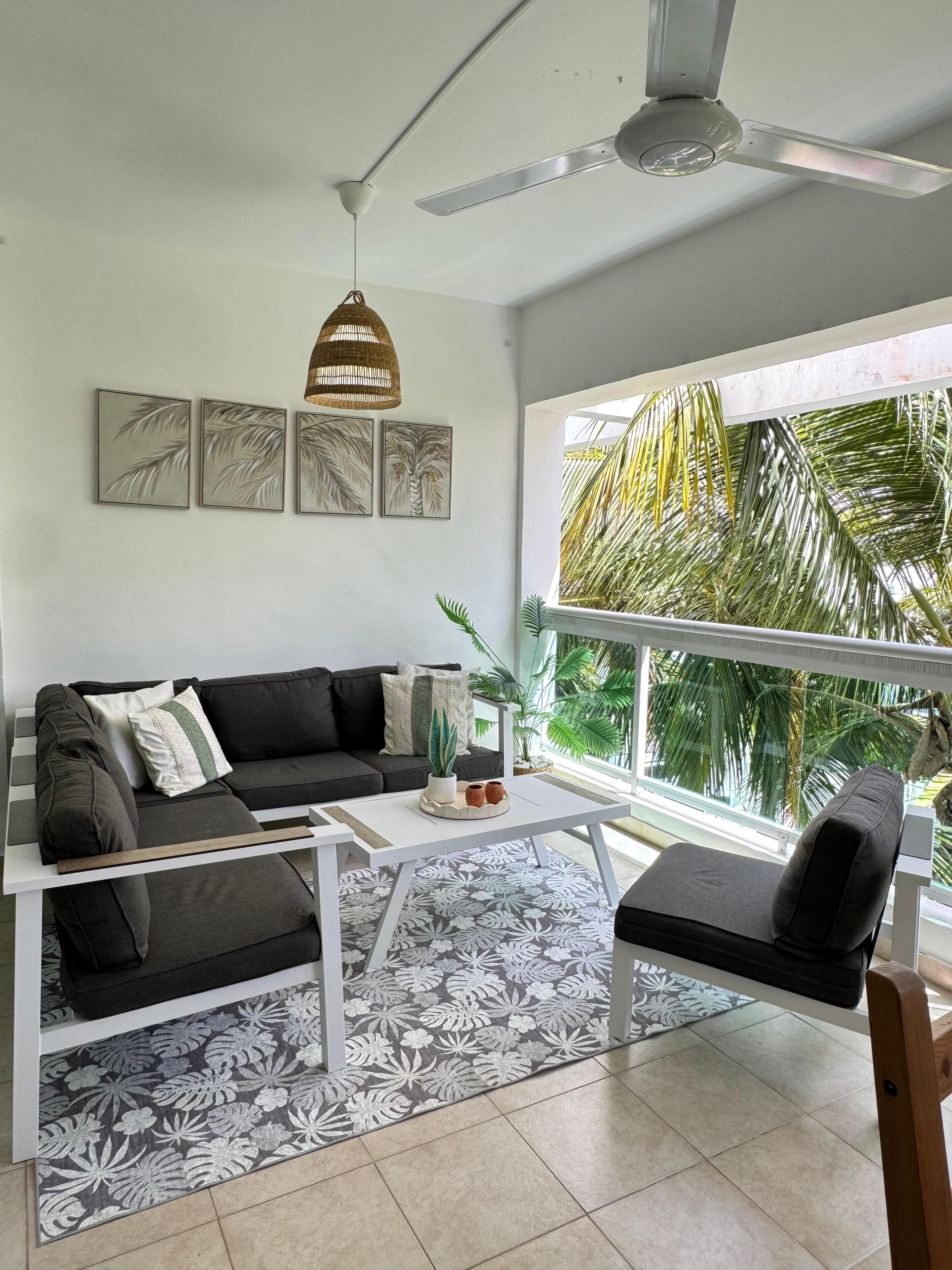 A modern living room with a sofa, coffee table, and a view of palm trees outside.