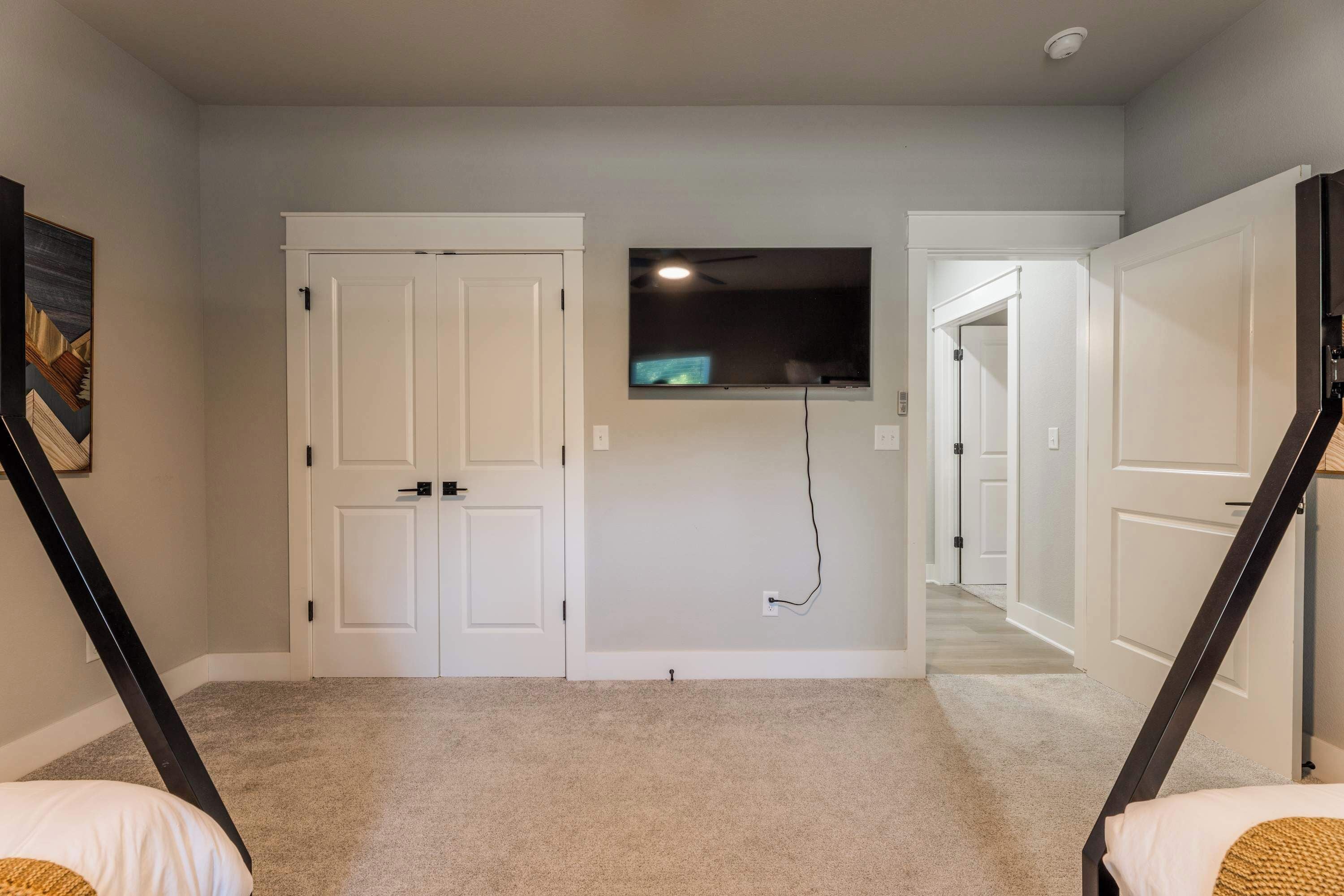 A room with a carpeted floor, two white doors, a mounted flat-screen TV, and a ceiling fan.