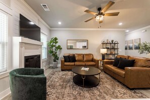 A modern living room with a fireplace, leather sofas, a coffee table, and a ceiling fan. - The Veloshire | Hot Tub + Back 40 Access (Bella Vista)