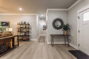 A hallway with a wooden floor, a black metal shelf with decorative items, a round mirror, and a door. - The Veloshire | Hot Tub + Back 40 Access (Bella Vista)