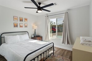 Una camera da letto con un letto, una ventola a soffitto, una finestra con tende, un comodino e quadri appesi al muro.