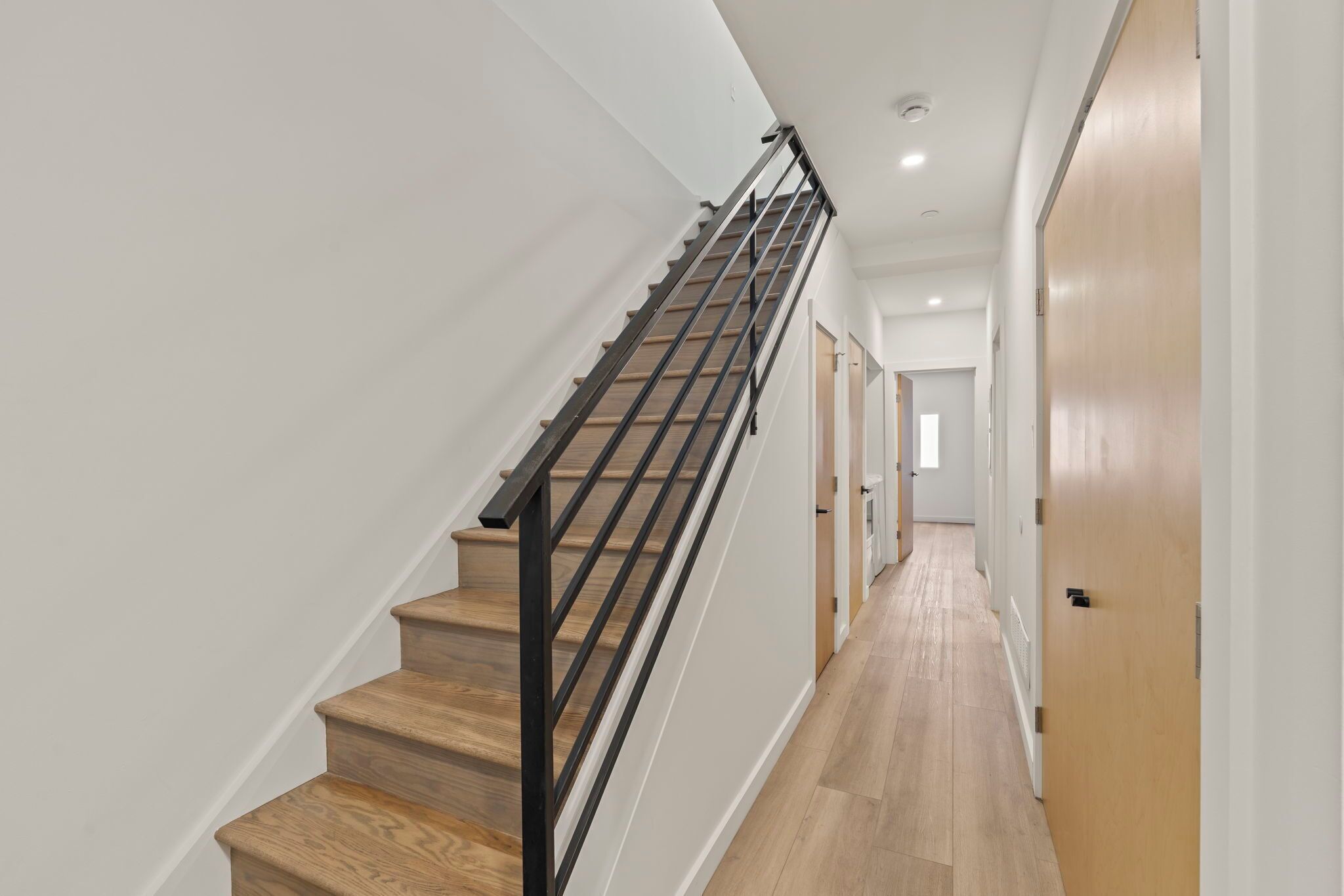 Una escalera moderna con escalones de madera y barandales negros, que conduce a un pasillo con paredes de tonos claros y suelo de madera.