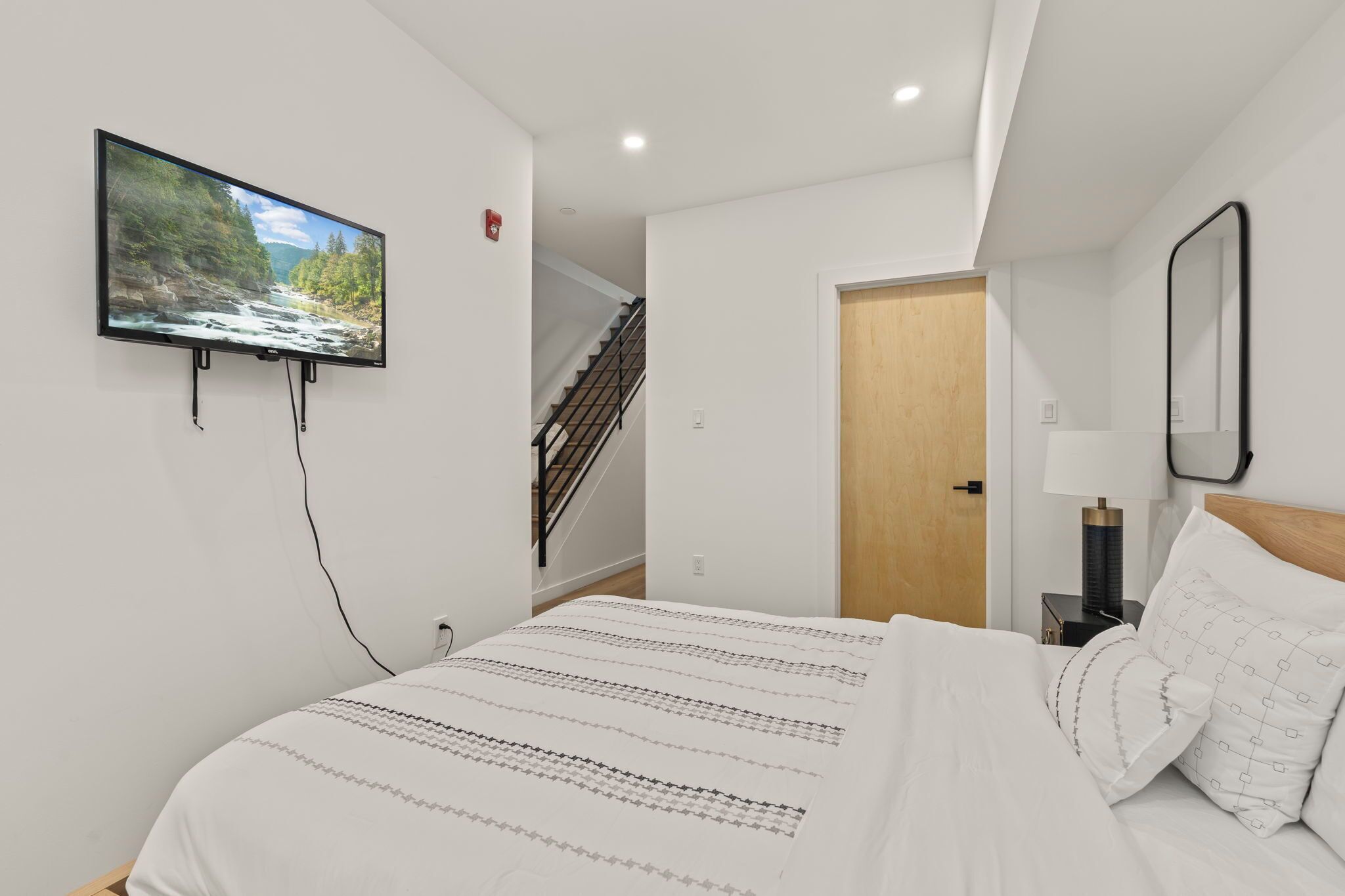 Ein Schlafzimmer mit einem Bett, einem an der Wand befestigten Fernseher, einem Nachttisch und einer Tür zu einem anderen Raum.