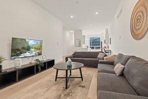 A modern living room with a flat-screen TV, a sofa, a coffee table, and a dining area in the background.