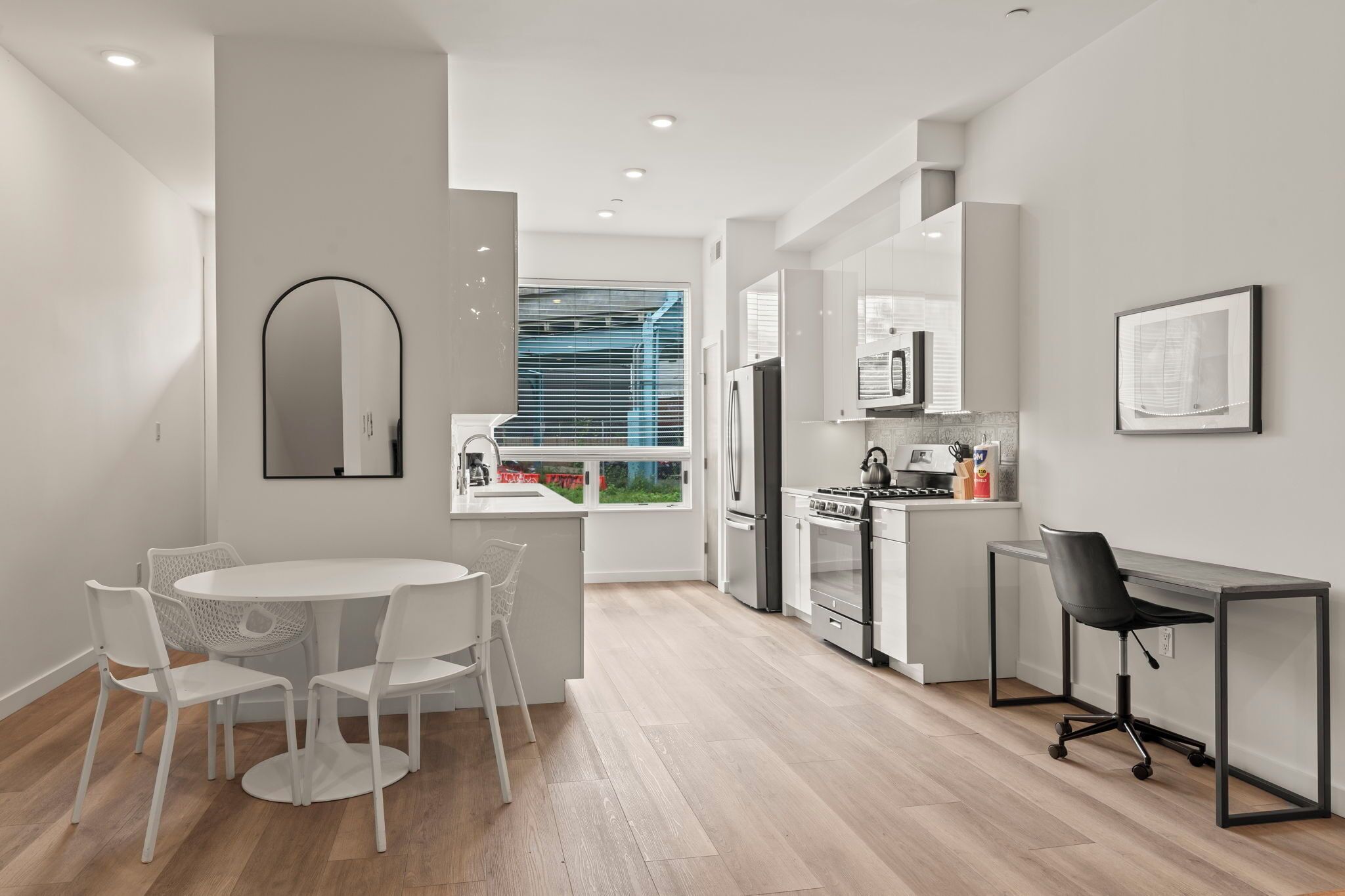 Una cocina moderna con armarios blancos, una estufa y un refrigerador. Hay un comedor con una mesa y sillas blancas, y un pequeño espacio de oficina con un escritorio y una silla.