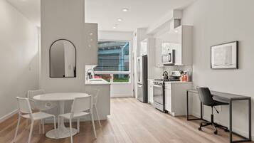 Una cocina moderna con armarios blancos, una estufa y un refrigerador. Hay un comedor con una mesa y sillas blancas, y un pequeño espacio de oficina con un escritorio y una silla.