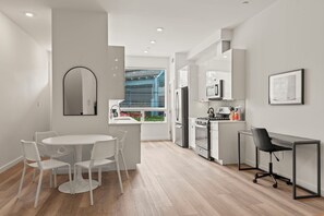 A modern kitchen with white cabinets, a stove, and a refrigerator. There is a dining area with a white table and chairs, and a small office space with a desk and chair.