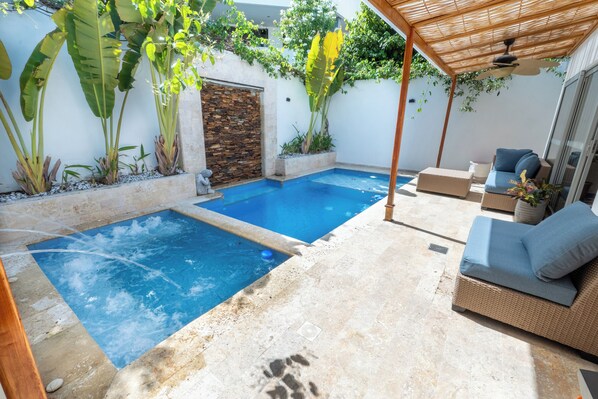 Ein Pool mit Steinmauer, umgeben von Pflanzen und einem überdachten Sitzbereich mit einer Couch.