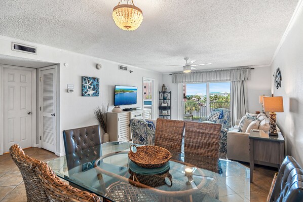 A spacious living room with a glass coffee table, wicker furniture, a television, and a view of the outdoors.