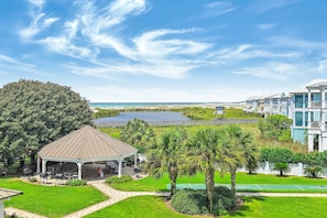 A gazebo with a roof, surrounded by a well-maintained lawn and palm trees, with a view of a beach and ocean in the background.