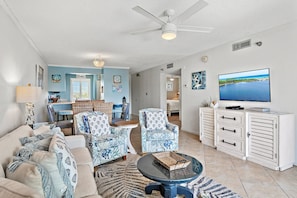 A living room with a sofa, two patterned armchairs, a coffee table, a TV on a cabinet, and a ceiling fan.