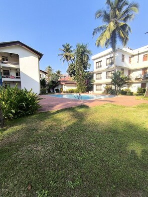 A pool area with a grassy foreground, palm trees, and multi-story buildings with balconies. - Welcome to Su Casa, your serene escape 600m away from the sun-kissed shores (Candolim)