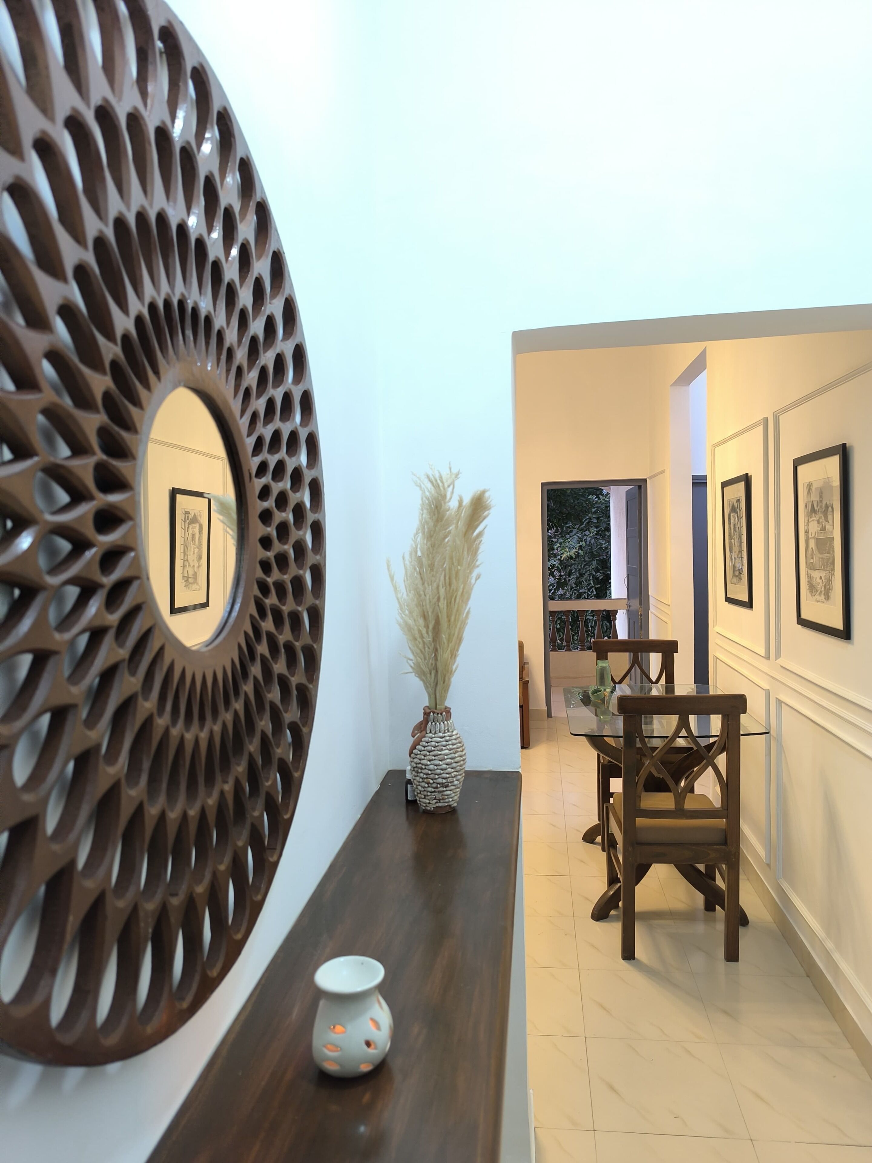 A hallway with a decorative wall piece, a vase with pampas grass, a glass table with chairs, and framed pictures on the wall.