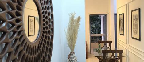 A hallway with a decorative wall piece, a vase with pampas grass, a glass table with chairs, and framed pictures on the wall.