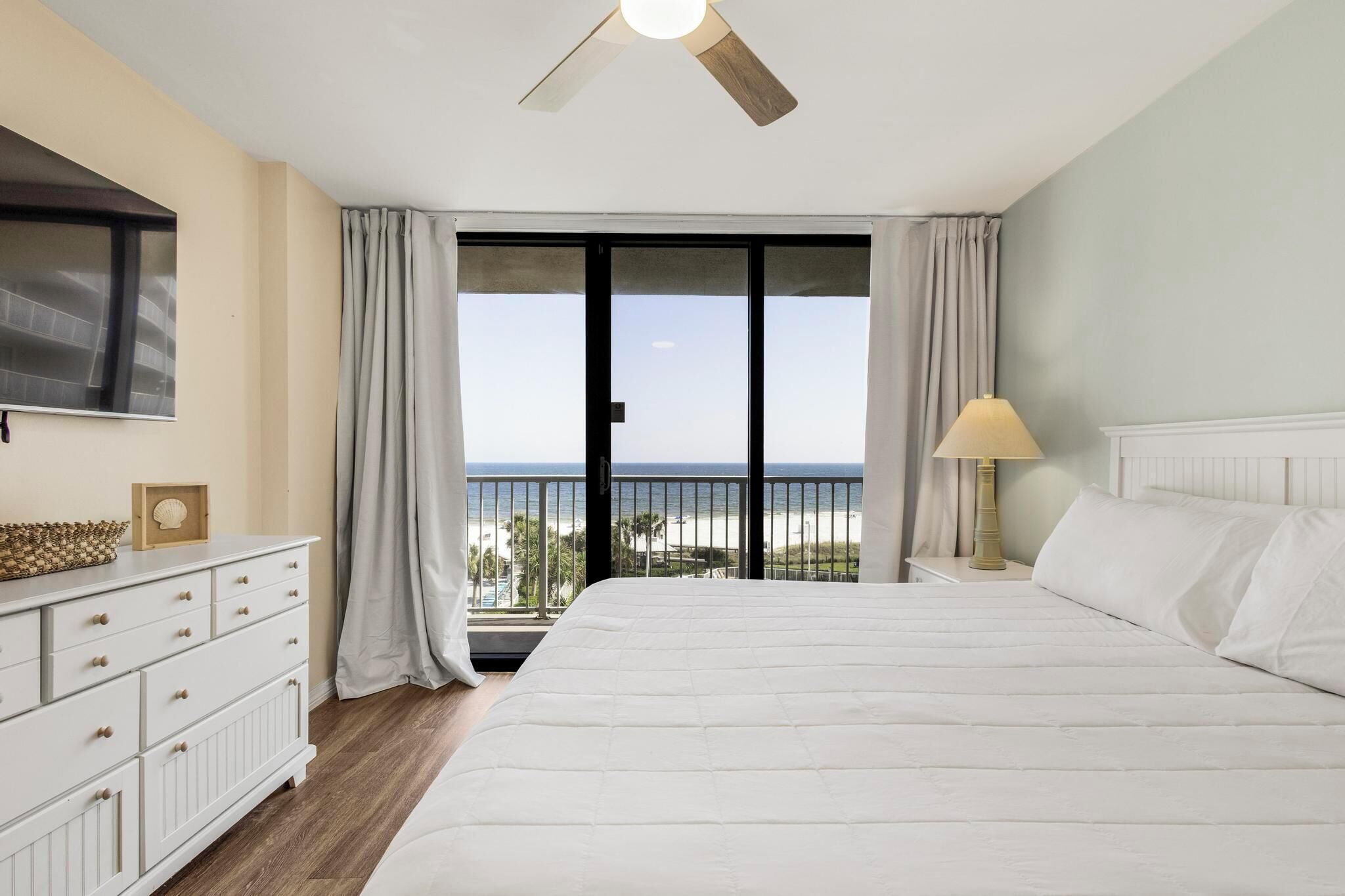 A hotel room with a large bed, a TV mounted on the wall, a bedside table with a lamp, and a view of the beach through the balcony doors.