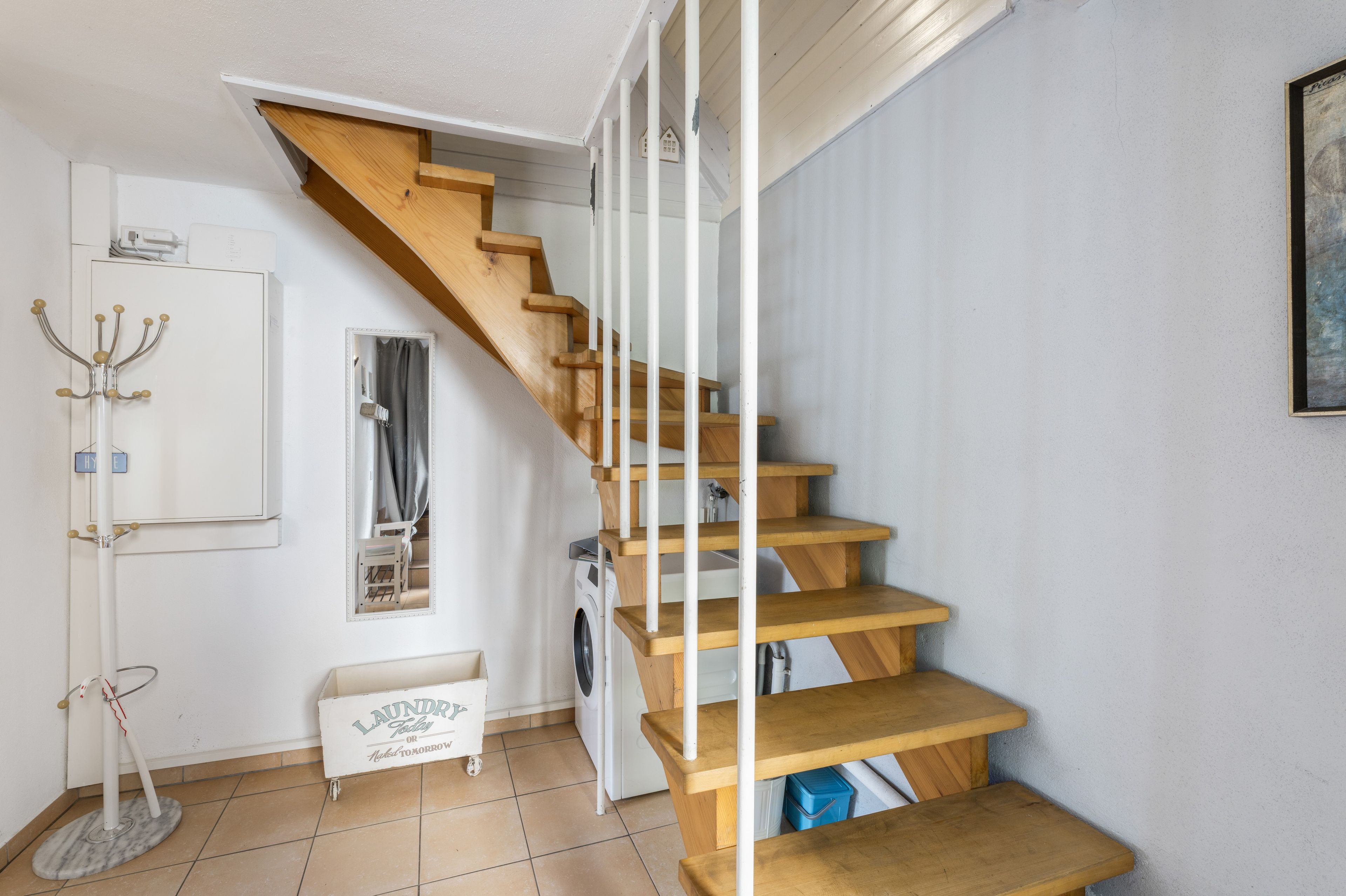 Una escalera con escalones de madera y barandillas blancas, una cesta de ropa, un perchero y una lavadora.
