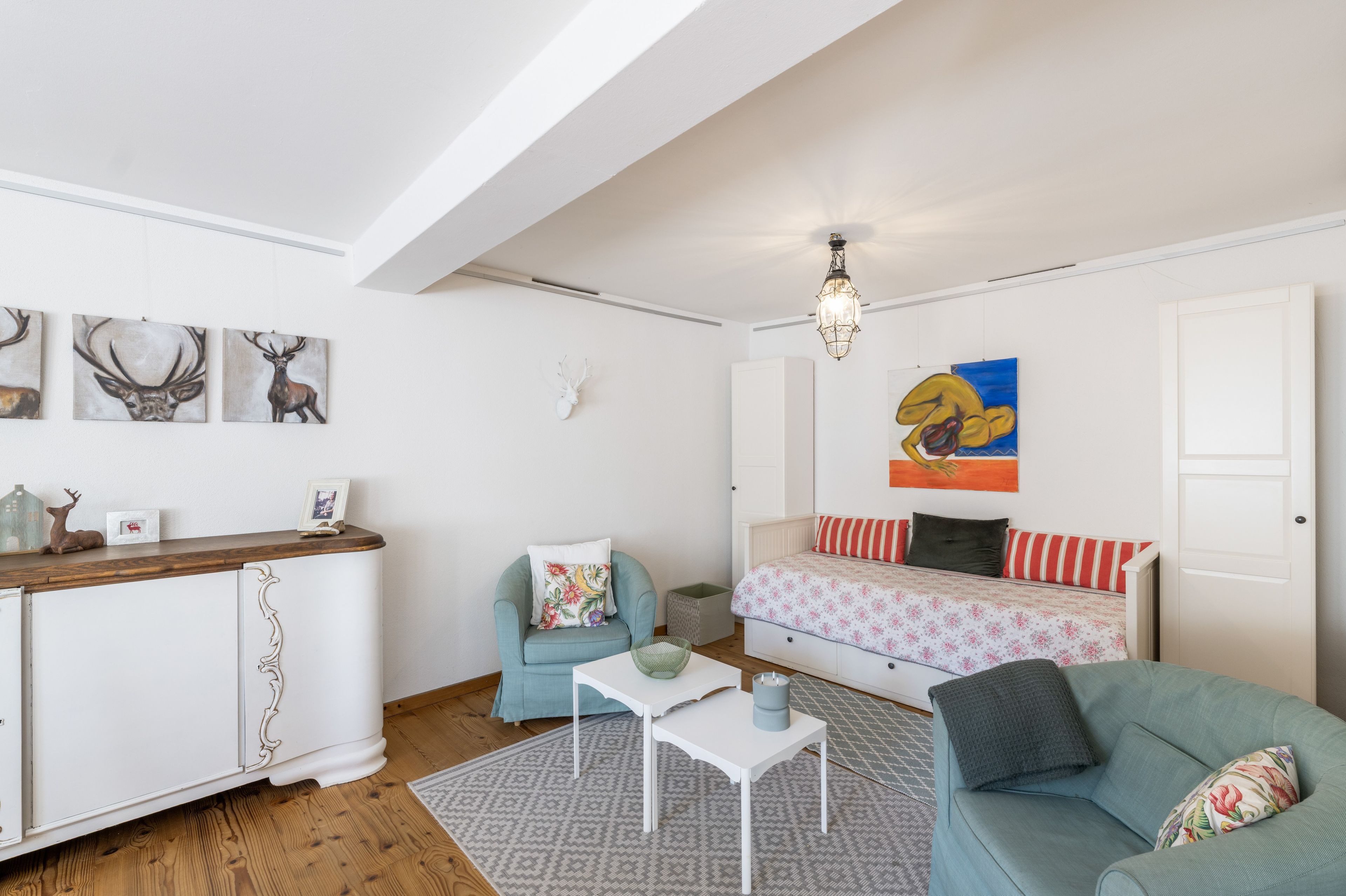 Ein Zimmer mit einer Couch, einem kleinen Tisch, einem Schrank und Wandbildern, darunter Darstellungen von Hirschen und einem Vogelgemälde.