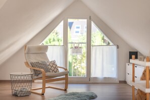 Een kamer met een schuine kap, een houten stoel met kussen, een klein rond tafeltje, een draadmand en een witte dressoir.