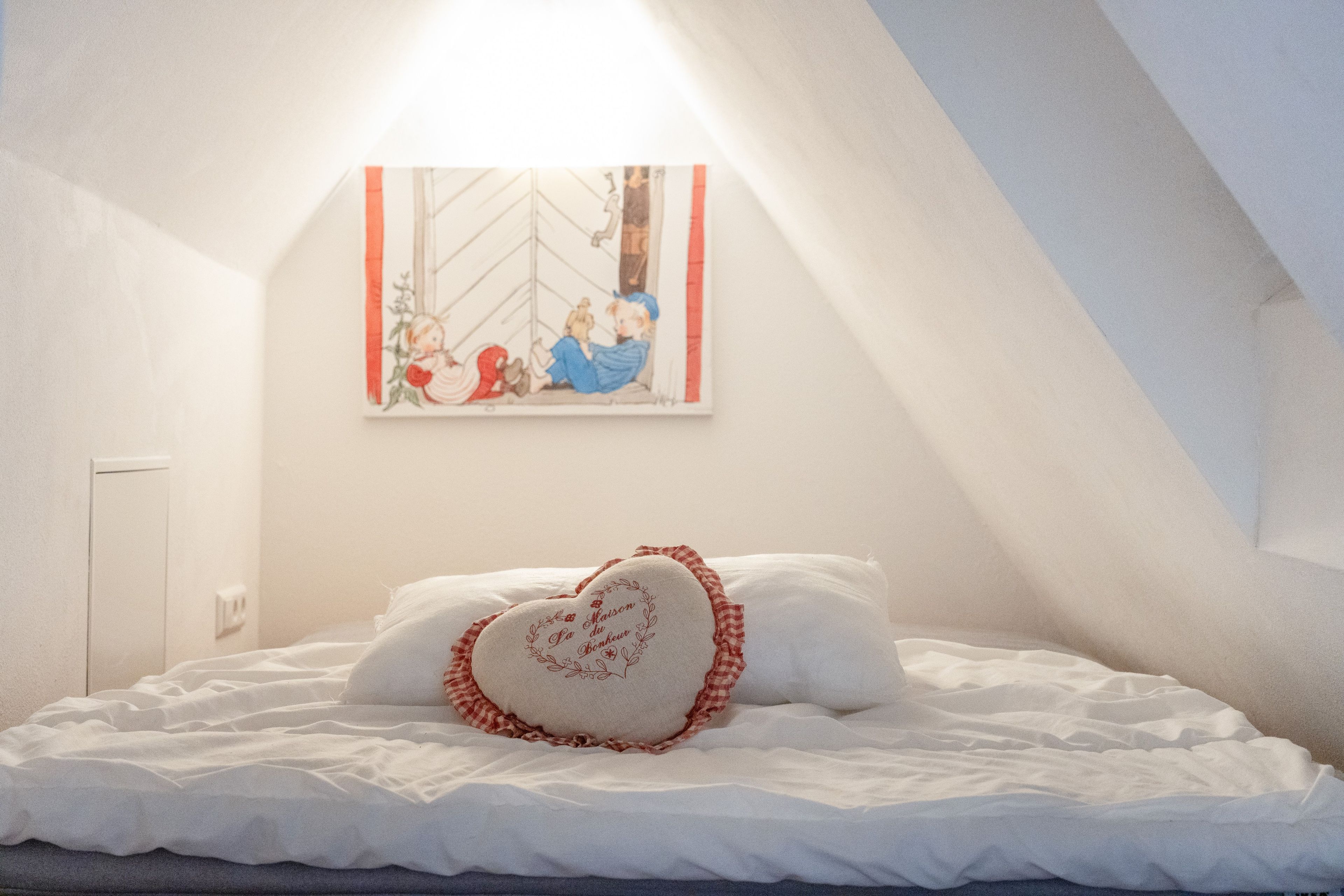 A bedroom with a bed, pillows, a heart-shaped pillow, a wall-mounted picture, and a sloped ceiling.